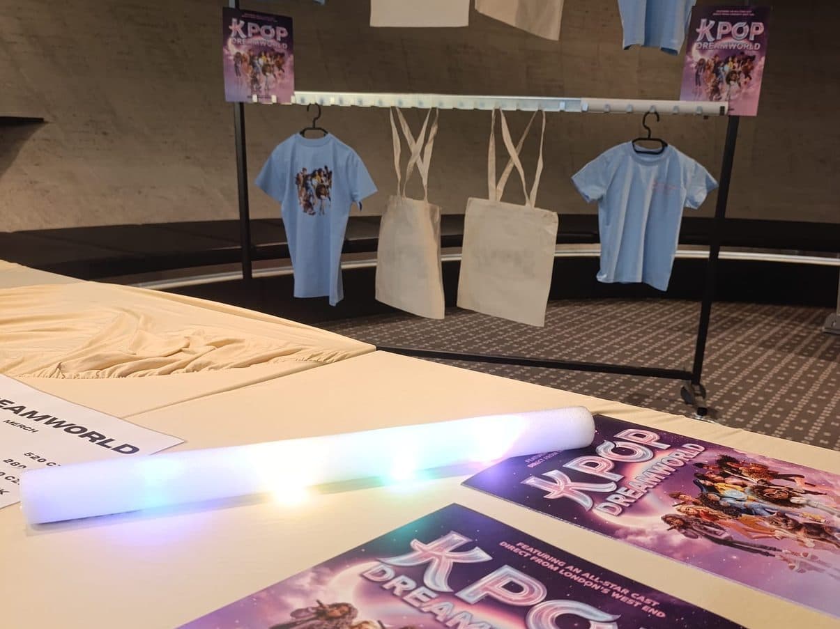 Prodejní stůl merch stánku turné K-POP DREAMWORLD s ceníkem a plakáty, za ním stojan s tričky a taškami